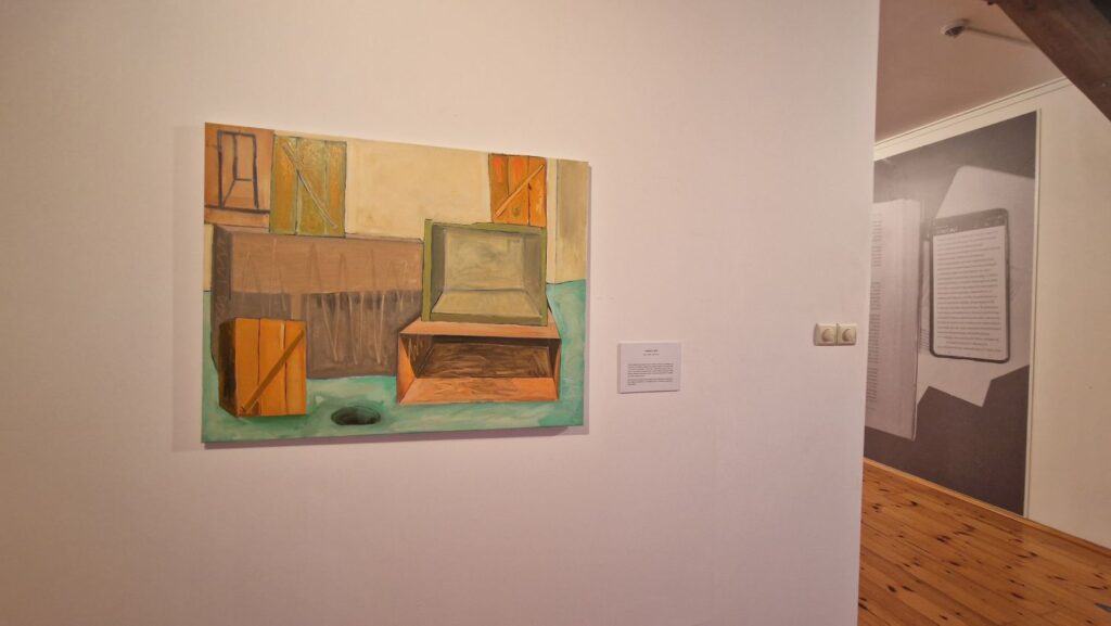 Obraz malarski przedstawiający uproszczone wnętrze z meblem przypominającym skrzynię lub łóżko, zawieszony na białej ścianie sali wystawowej (kliknięcie powiększy zdjęcie)