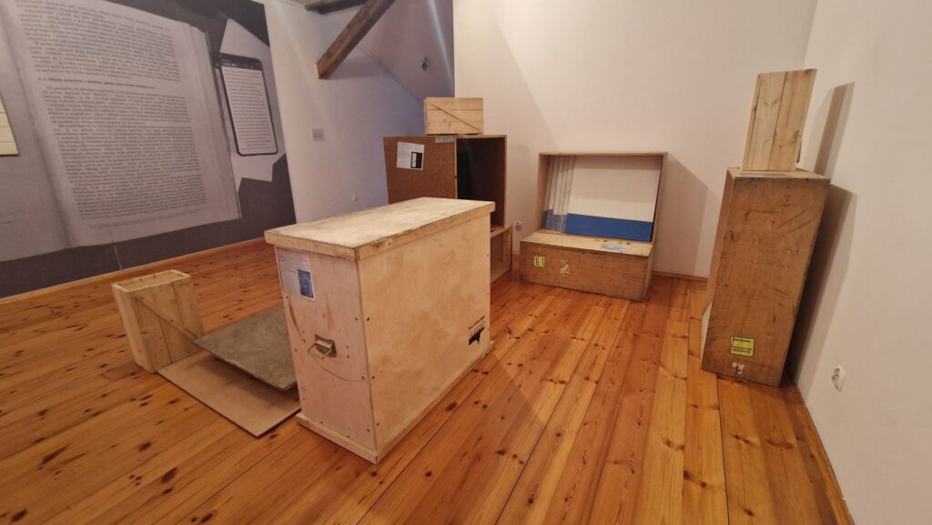 Instalacja artystyczna złożona z drewnianych skrzyń i otwartych pojemników ustawionych na podłodze; w tle fragmenty plansz z tekstami towarzyszącymi wystawie (kliknięcie powiększy zdjęcie)