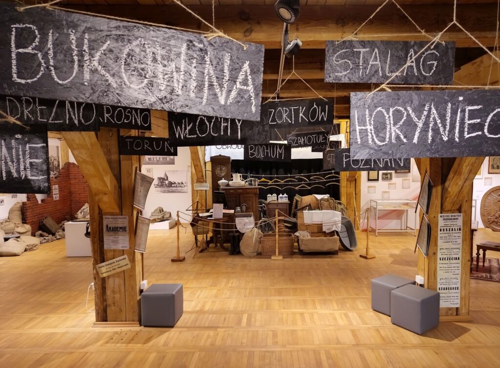 Wnętrze sali muzealnej z aranżacją wystawy historycznej, nad którą wiszą tablice z nazwami miejscowości zapisanymi kredą, a w centrum znajdują się meble, walizki i eksponaty z okresu powojennego (kliknięcie powiększy zdjęcie).