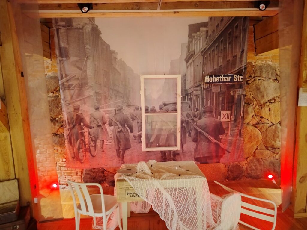 Aranżacja wystawy historycznej z dużą fotografią zniszczonego miasta i żołnierzy na ulicy, przed którą ustawiono stół przykryty koronkową tkaniną oraz krzesła, a całość oświetlona jest czerwonym światłem (kliknięcie powiększy zdjęcie).