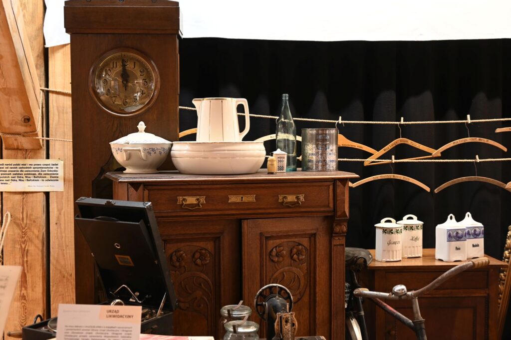 Drewniana komoda z zegarem i naczyniami, obok stojący rower oraz elementy wyposażenia domowego tworzące aranżację wnętrza z okresu historycznego (kliknięcie powiększy zdjęcie).