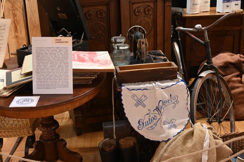 Zbliżenie na eksponaty: stolik z dokumentami, haftowaną serwetą i przedmiotami codziennego użytku, obok widoczny fragment roweru i wyposażenia wnętrza (kliknięcie powiększy zdjęcie).