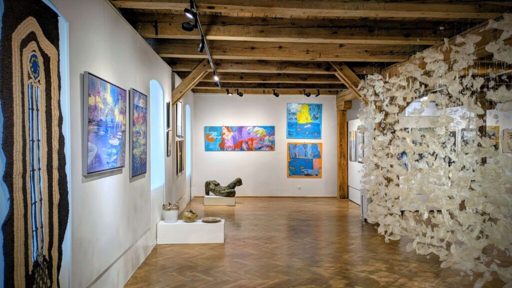 Widok sali wystawowej z podłużną tkaniną artystyczną i instalacją przestrzenną z białych elementów (kliknięcie powiększy zdjęcie).