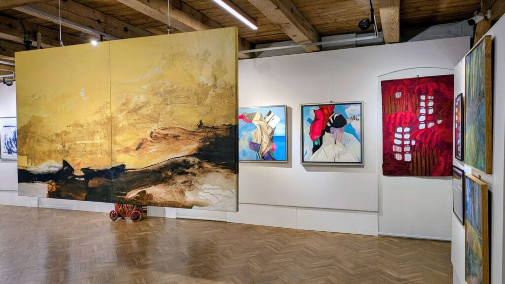 Duży obraz w odcieniach złota prezentowany centralnie na wystawie, w tle inne prace malarskie (kliknięcie powiększy zdjęcie).