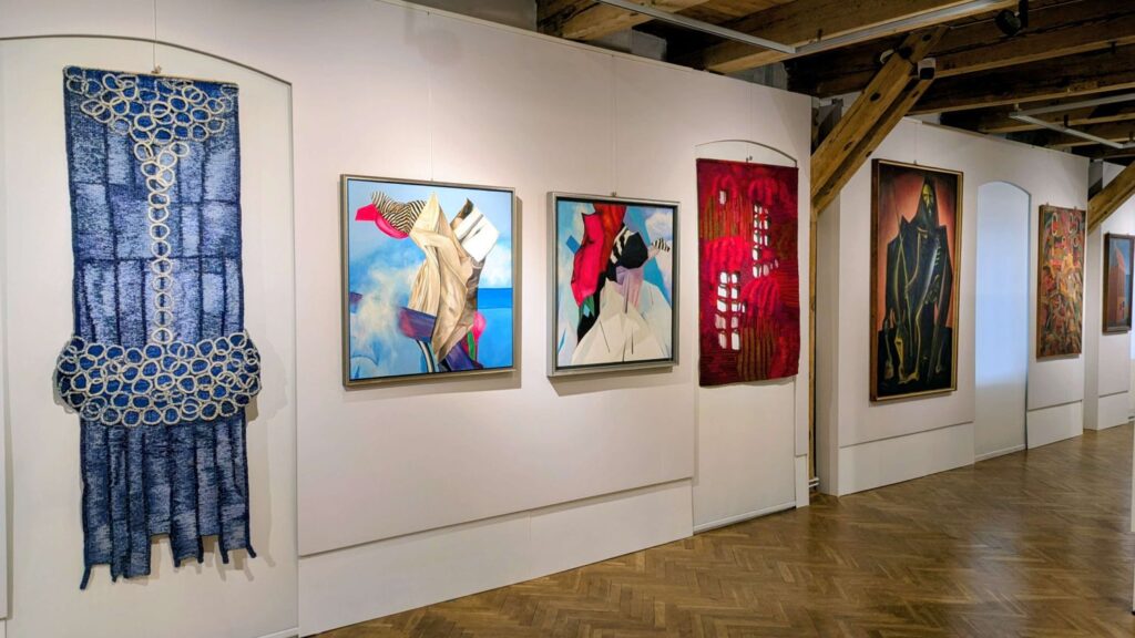 Sala wystawy z ekspozycją obrazów na białych ścianach i drewnianymi belkami stropowymi (kliknięcie powiększy zdjęcie).