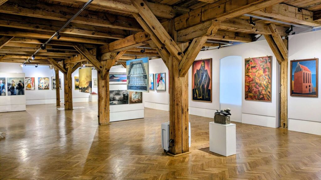 Przestronna sala wystawowa z pracami malarskimi i rzeźbą na kubiku ekspozycyjnym, widoczne drewniane słupy konstrukcyjne (kliknięcie powiększy zdjęcie).