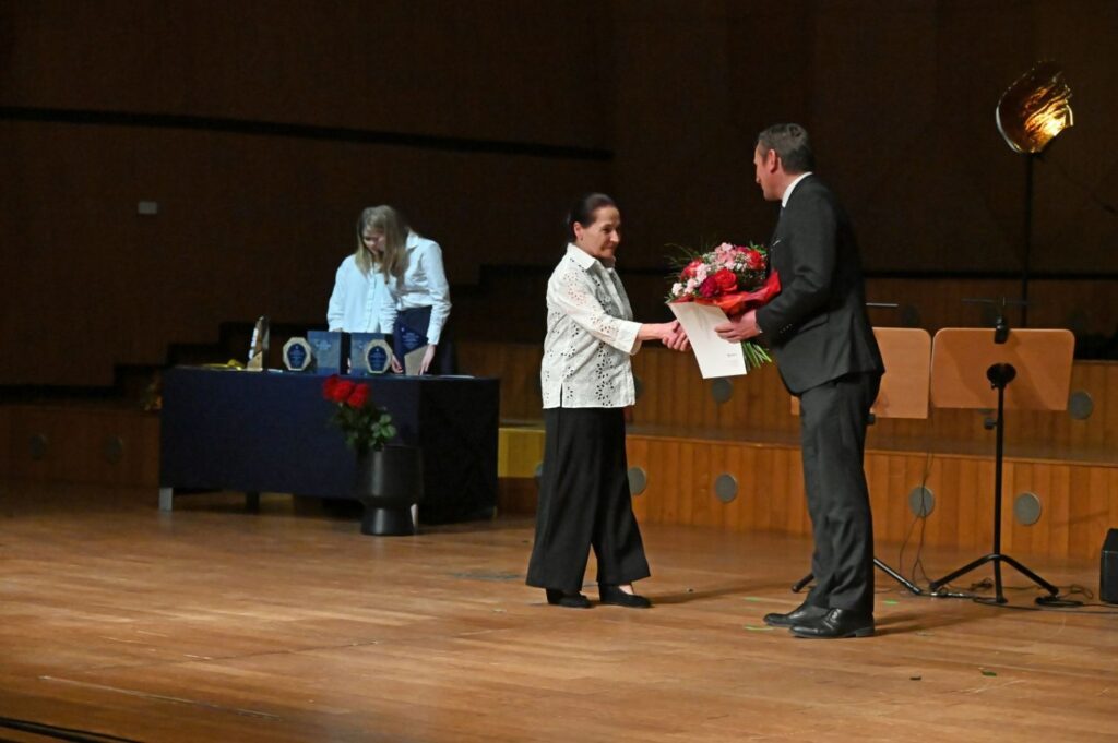 Kobieta odbiera bukiet kwiatów od mężczyzny na scenie podczas uroczystej gali (kliknięcie powiększy zdjęcie).