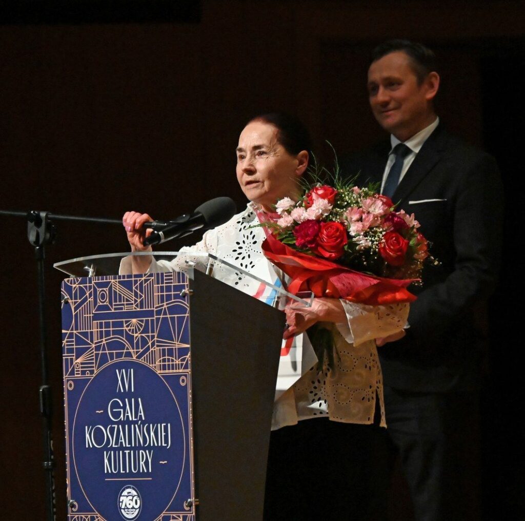 Walentyna Orłowska przemawia przy mównicy podczas gali, trzymając bukiet kwiatów, obok stoi mężczyzna w garniturze (kliknięcie powiększy zdjęcie).