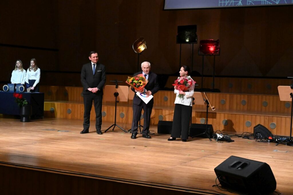 Dwóch laureatów stojących na scenie w towarzystwie organizatorów wydarzenia, jeden z nich trzyma w rękach kwiaty a drugi dyplom (kliknięcie powiększy zdjęcie).