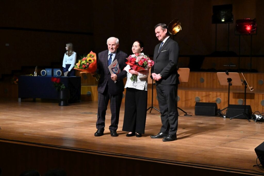 Dwóch nagrodzonych laureatów z bukietami i dyplomami pozuje do zdjęcia na scenie, wśród nich prezydent miasta (kliknięcie powiększy zdjęcie).