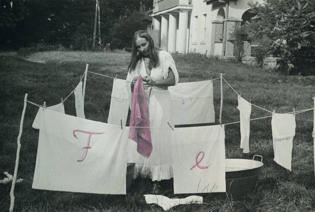 Czarno-biała fotografia performansu Marii Pinińskiej-Bereś z 1981 roku. Kobieta stoi przy rozwieszonym praniu na trawie przed budynkiem, trzymając różową tkaninę. Na białych płachtach widoczne są różowe litery. (kliknięcie powiększy zdjęcie).