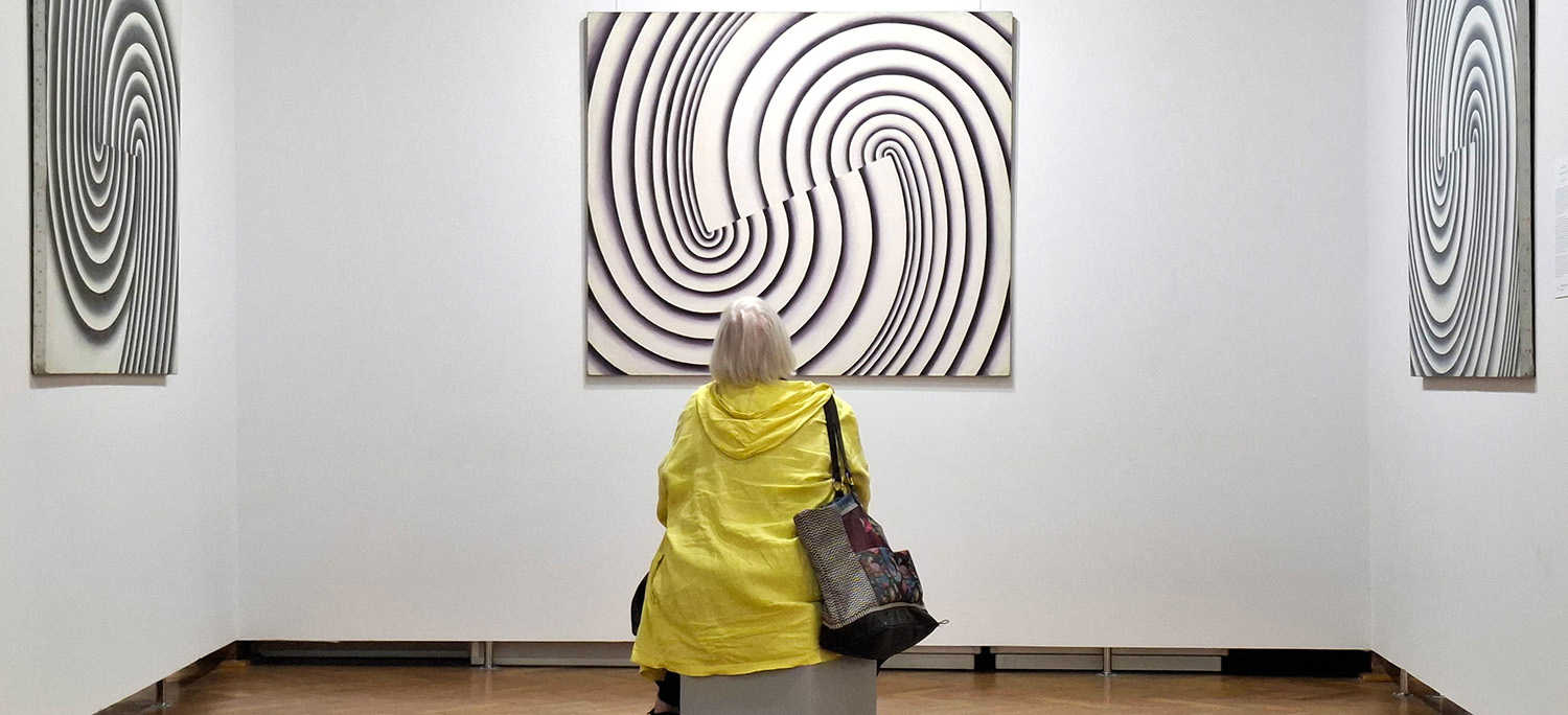 Zwiedzająca siedzi na ławce i ogląda abstrakcyjne reliefy prezentowane na wystawie sztuki współczesnej. Trzy kompozycje o spiralnych i falujących liniach wiszą na białych ścianach galerii.