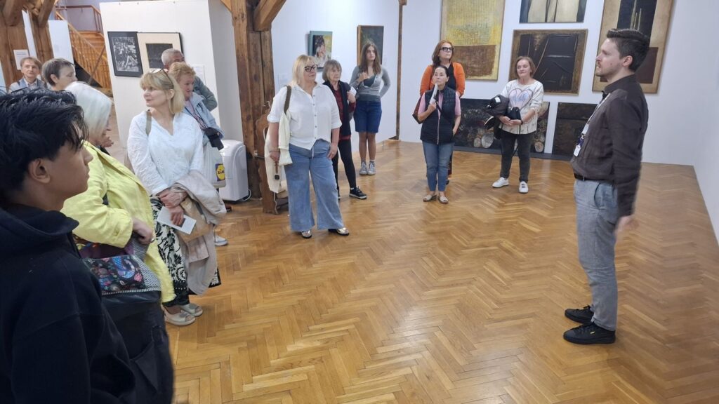 Grupa zwiedzających słucha komentarza kuratora wystawy w jednej z sal wystawowych, otoczona obrazami prezentowanymi na ekspozycji sztuki współczesnej (kliknięcie powiększy zdjęcie).