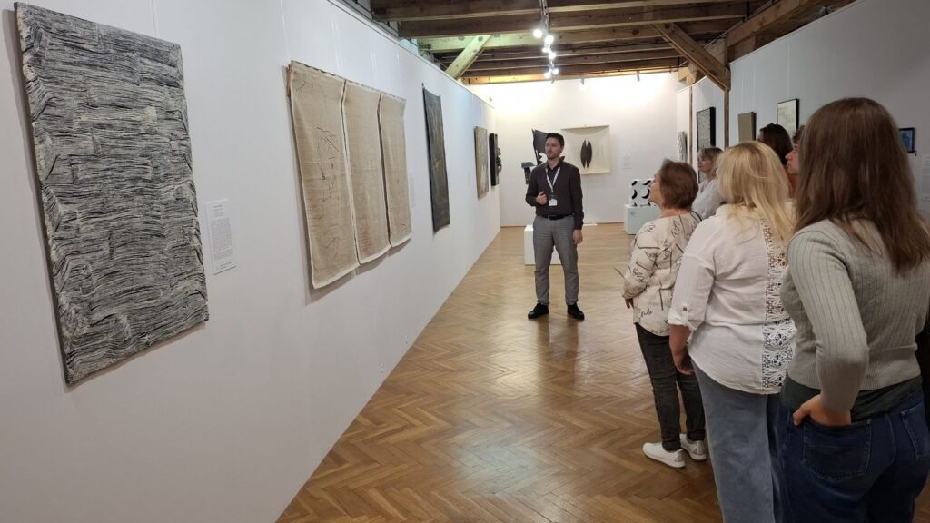 Grupa zwiedzających słucha opowieści kuratora podczas przejścia przez galerię z obrazami i instalacjami sztuki współczesnej (kliknięcie powiększy zdjęcie).