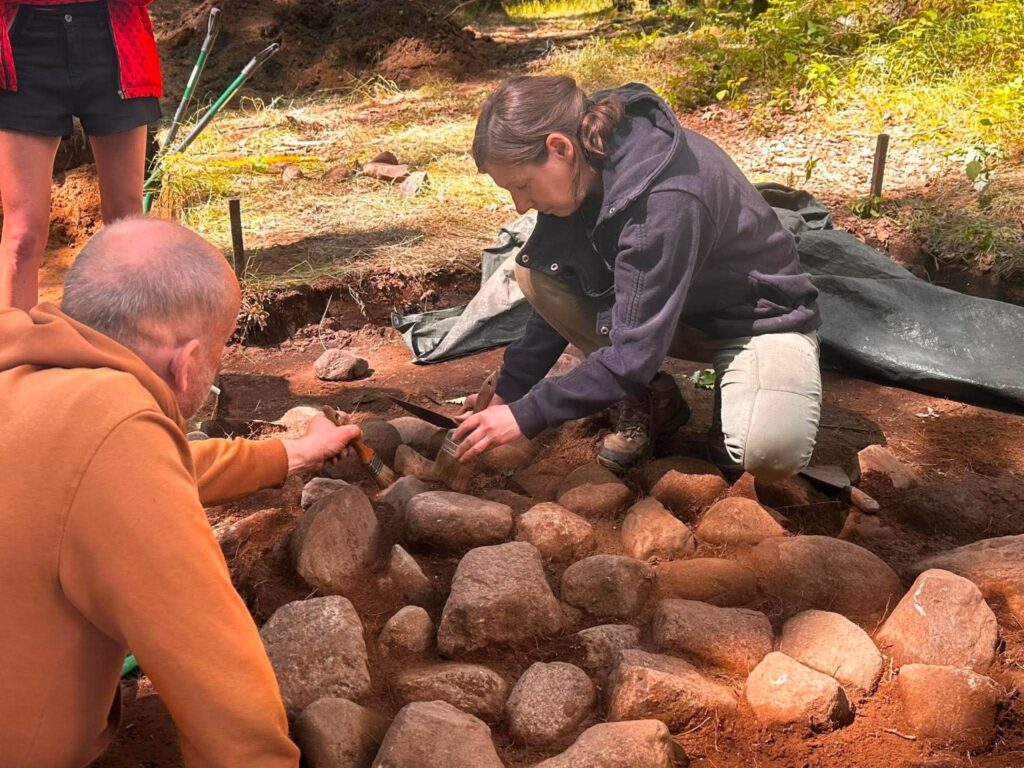 Osoba będąca w wykopie oczyszcza odsłoniętą kamienną konstrukcję, usuwając ziemię spomiędzy otoczaków. Obok niej druga osoba przygląda się postępom prac i pomaga w dokumentowaniu odkrycia (kliknięcie powiększy zdjęcie).