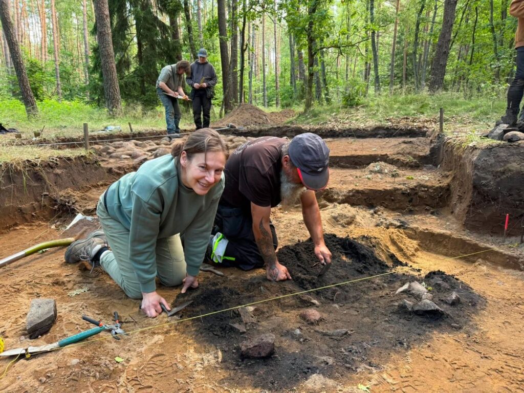 Dwie osoby pracują przy oczyszczaniu ciemnych pozostałości odkrytych w wykopie archeologicznym. W tle widoczne są kolejne wykopy oraz uczestnicy badań dokumentujący odkrycia na leśnym stanowisku (kliknięcie powiększy zdjęcie).