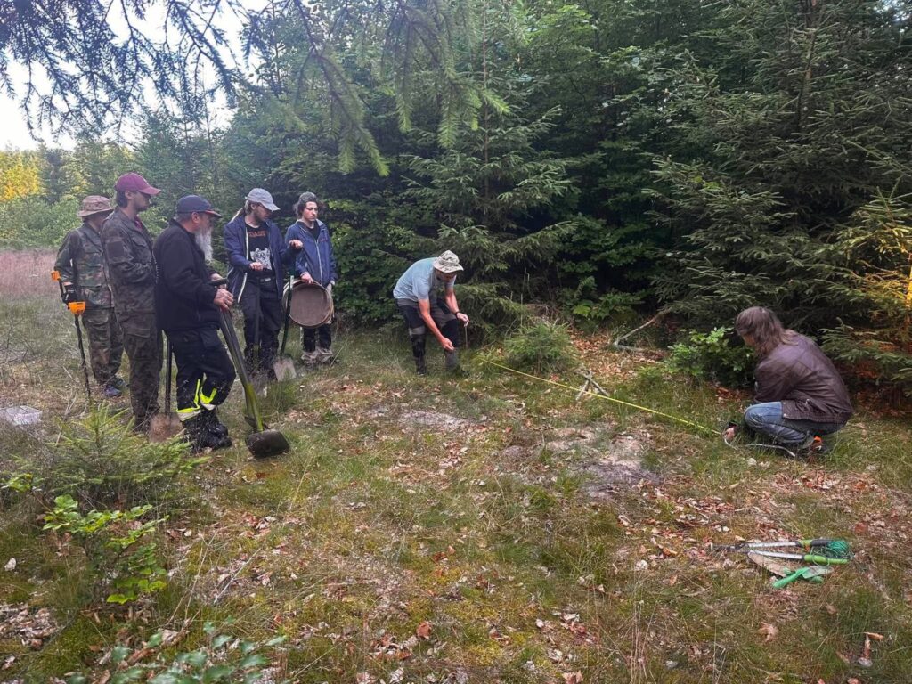 Grupa archeologów, studentów i wolontariuszy przygotowuje stanowisko badawcze w lesie. Część uczestników obserwuje wyznaczanie granic wykopu przy użyciu taśmy mierniczej, a w tle widoczne są gęste drzewa (kliknięcie powiększy zdjęcie).