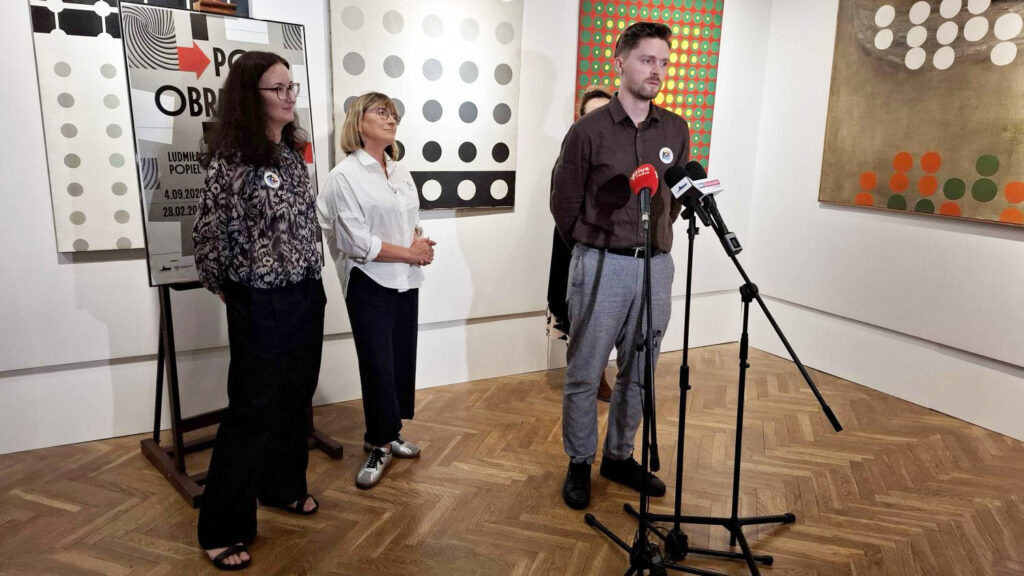 Trzy osoby stoją przed mikrofonami podczas konferencji prasowej na sali wystawowej; w tle widoczne są abstrakcyjne obrazy oraz plansza informacyjna wystawy (kliknięcie powiększy zdjęcie).