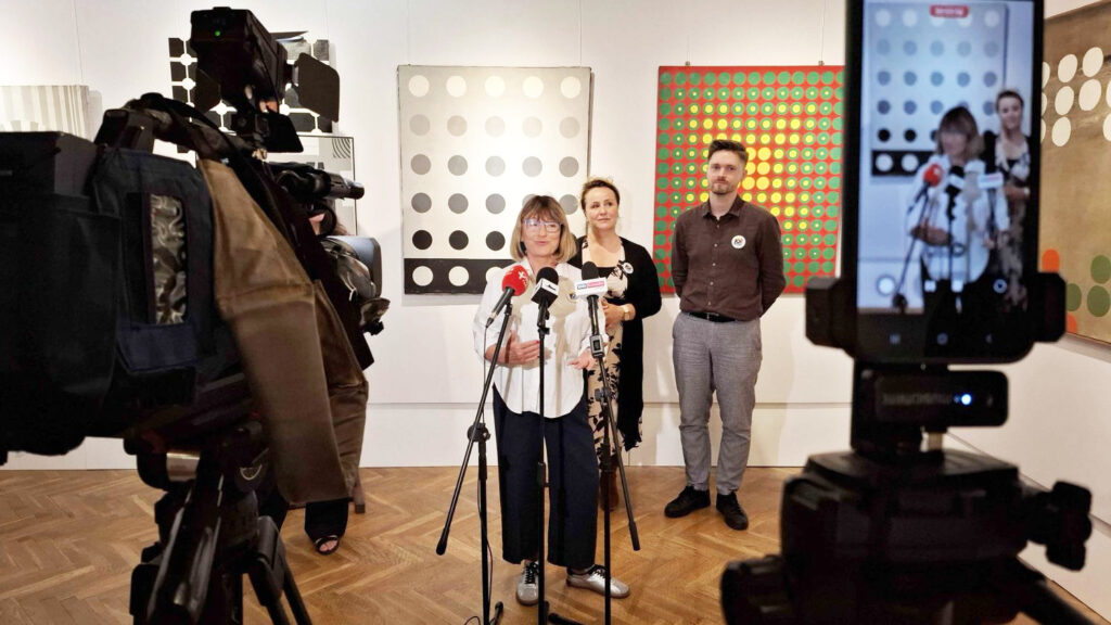 Kamera telewizyjna rejestruje wypowiedź kobiety stojącej przy mikrofonach; za nią stoją pozostali uczestnicy konferencji prasowej; na pierwszym planie kamera telewizyjna i telefon na statywie, który nagrywa wydarzenie (kliknięcie powiększy zdjęcie).