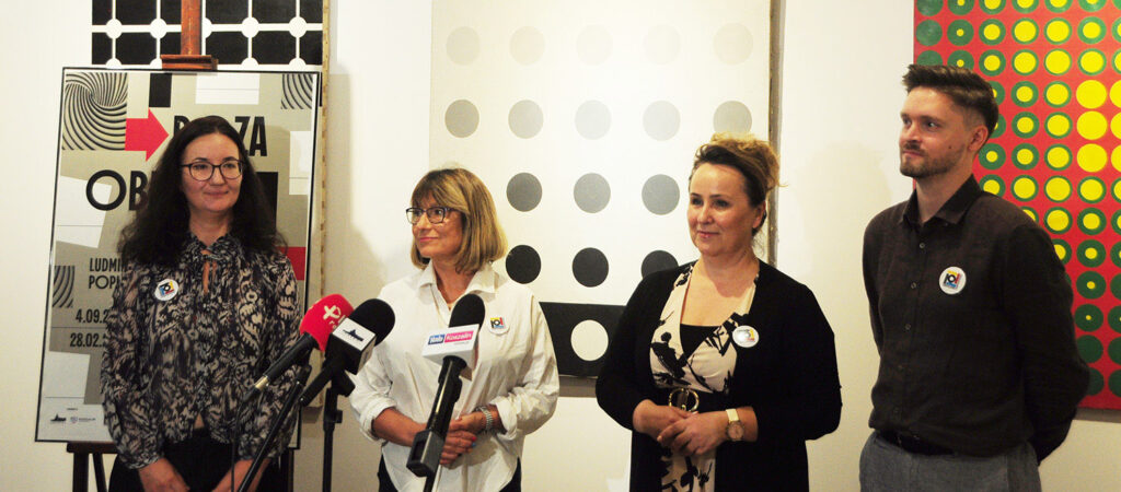 Cztery osoby stoją przed mikrofonami i czekają na rozpoczęcie konferencji prasowej; w tle widoczne są abstrakcyjne obrazy oraz plansza informacyjna wystawy (kliknięcie powiększy zdjęcie).