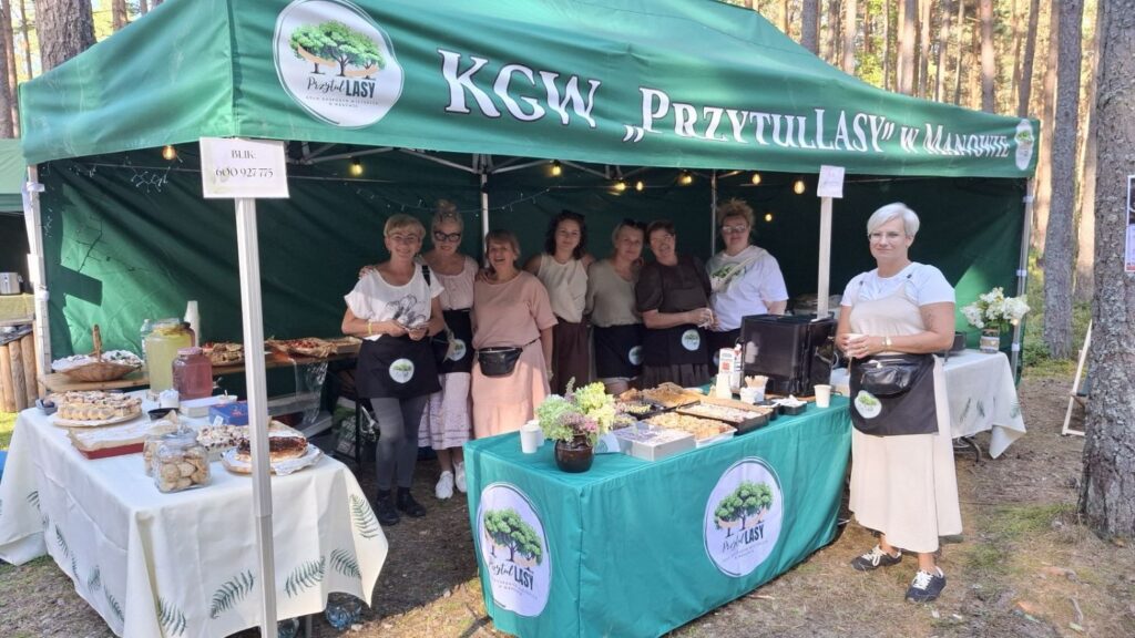 Grupa kobiet prezentuje stoisko Koła Gospodyń Wiejskich w lesie; na stołach znajdują się domowe wypieki i inne produkty przygotowane na wydarzenie (kliknięcie powiększy zdjęcie).