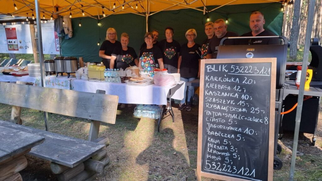 Stoisko gastronomiczne pod zadaszeniem; za ladą stoi grupa osób, a przed nim znajduje się tablica z odręcznie zapisanym menu i cenami potraw (kliknięcie powiększy zdjęcie).