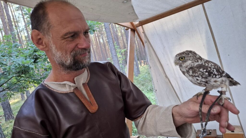 Mężczyzna w historycznym stroju trzyma na dłoni sowę; obok widoczny jest namiot stanowiska edukacyjnego (kliknięcie powiększy zdjęcie).