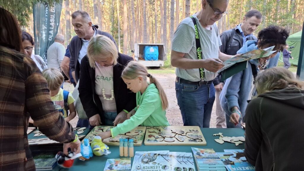 Dzieci i dorośli uczestniczą w zajęciach przy stoisku edukacyjnym; na stole przygotowano materiały do zabaw i aktywności dla odwiedzających (kliknięcie powiększy zdjęcie).