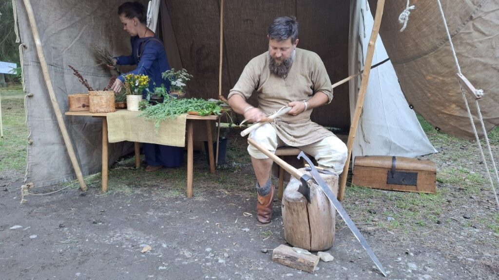 Mężczyzna w historycznym stroju wykonuje ręczne prace przy drewnianym stole; obok znajdują się narzędzia, materiały i pojemniki z roślinami (kliknięcie powiększy zdjęcie).