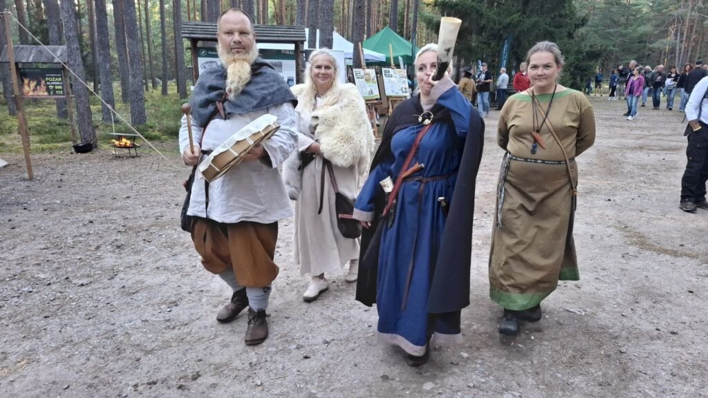 Grupa osób w strojach inspirowanych epoką średniowiecza spaceruje leśną ścieżką; uczestnicy trzymają historyczne rekwizyty i rozmawiają z odwiedzającymi (kliknięcie powiększy zdjęcie).