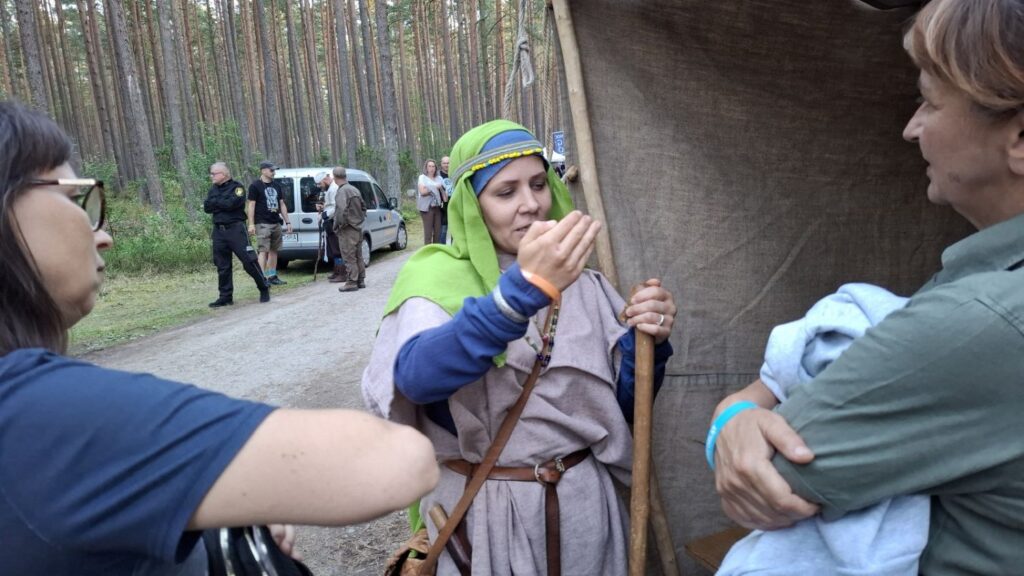 Kobieta w historycznym stroju rozmawia z uczestnikami wydarzenia; obok stoi osoba z mikrofonem, a spotkanie odbywa się przy jednym ze stanowisk w lesie (kliknięcie powiększy zdjęcie).