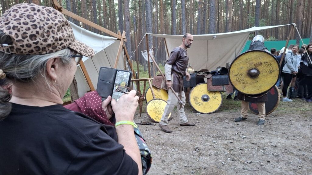 Uczestnicy obserwują pokaz z użyciem tarcz i historycznego uzbrojenia; na pierwszym planie jedna z osób fotografuje wydarzenie telefonem (kliknięcie powiększy zdjęcie).