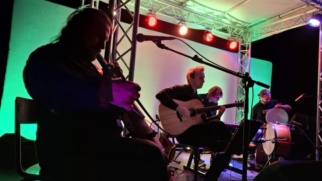 Muzycy grają na instrumentach smyczkowych, gitarze i instrumentach perkusyjnych. Scenę oświetlają czerwone i zielone reflektory (kliknięcie powiększy zdjęcie).