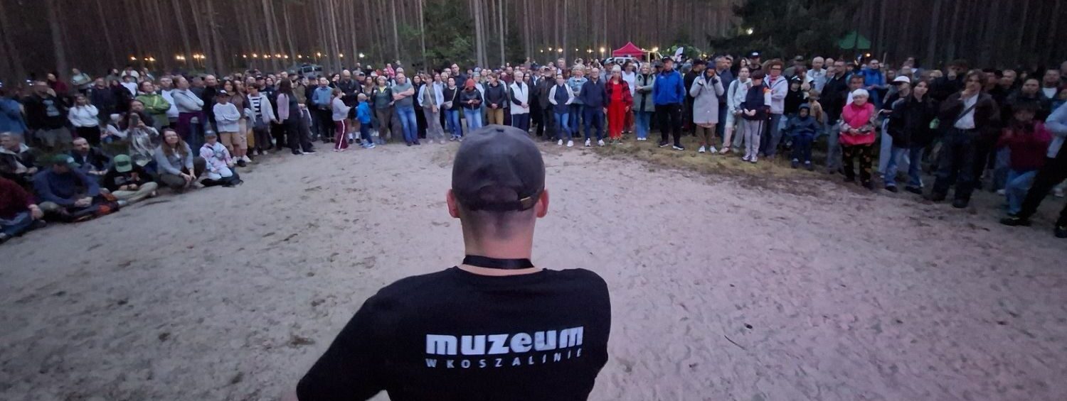 Archeolog z muzeum stoi tyłem do kadru, na piaszczystym terenie; przed nim stoją setki uczestników wydarzenia, którzy słuchają wprowadzenia do oprowadzania po rezerwacie; w tle drzewa i rozwieszone na nich oświetlenie.