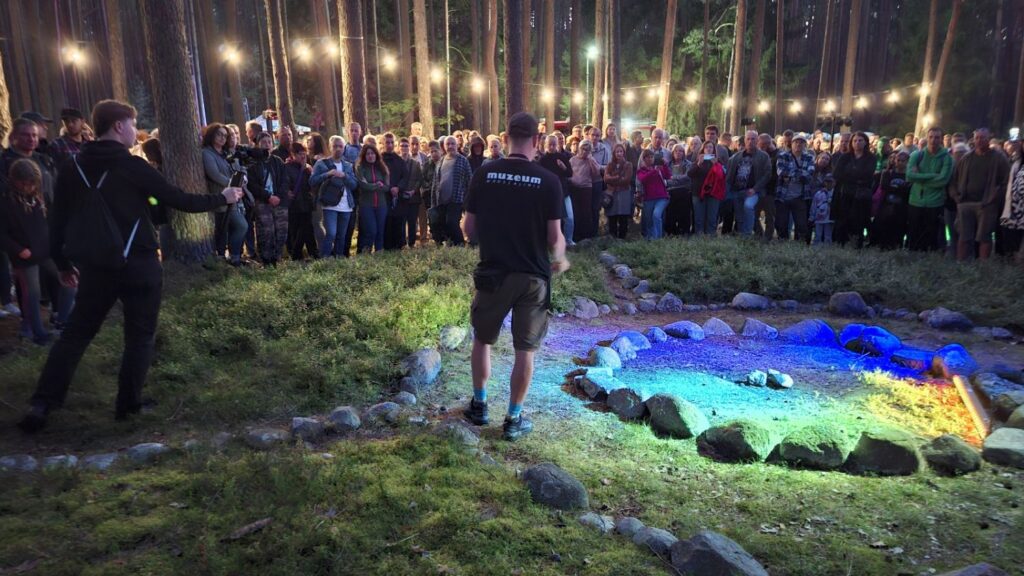 Grupa uczestników obserwuje podświetlony kamienny krąg; nad nimi, między drzewami, rozwieszone są punkty świetlne oświetlające miejsce wydarzenia (kliknięcie powiększy zdjęcie).