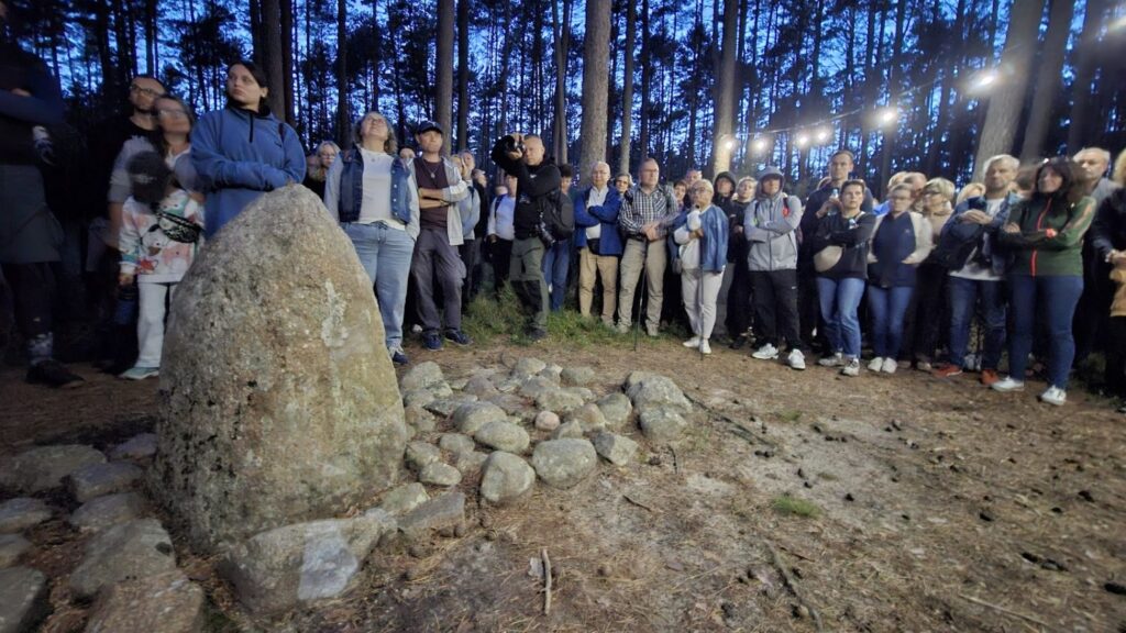 Duża grupa uczestników zgromadziła się wokół kamiennej instalacji z dużym głazem pośrodku; miejsce oświetlają lampy rozwieszone między drzewami (kliknięcie powiększy zdjęcie).
