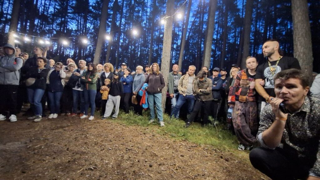 Liczna grupa osób stoi w lesie podczas nocnego wydarzenia; część uczestników obserwuje instalację, a na pierwszym planie mężczyzna przykuca z mikrofonem (kliknięcie powiększy zdjęcie).
