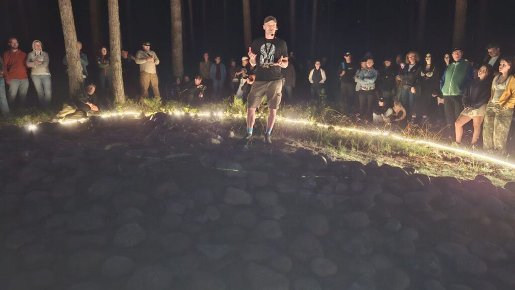 Mężczyzna stoi wewnątrz dużego kamiennego kręgu i zwraca się do zgromadzonych wokół uczestników; krąg jest podświetlony jasną linią światła (kliknięcie powiększy zdjęcie).