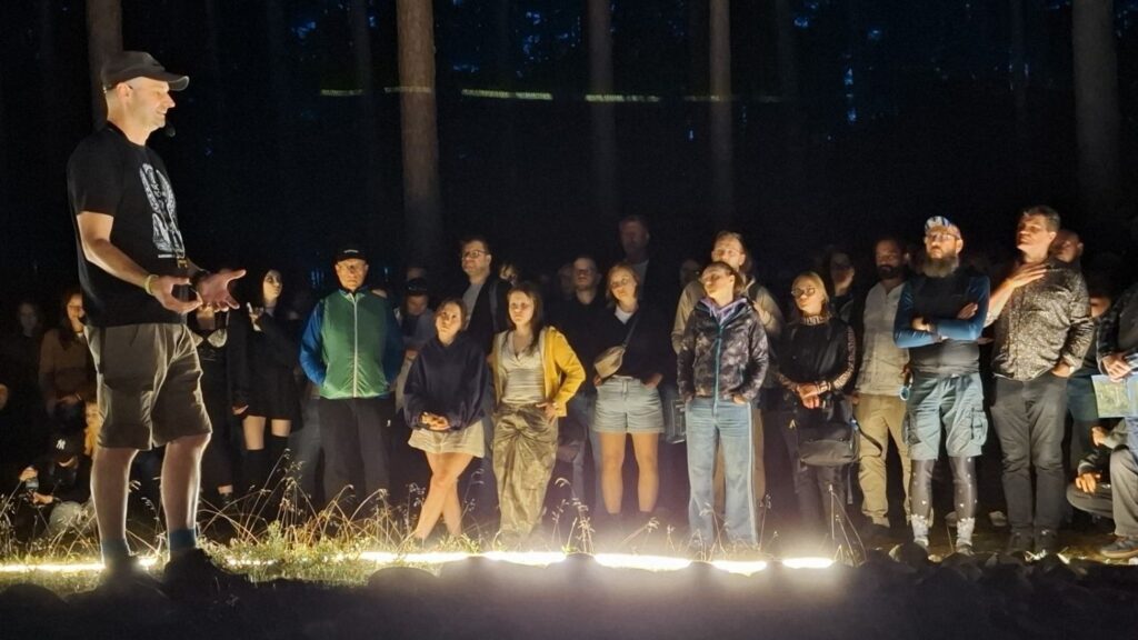 Mężczyzna stoi na skraju podświetlonego kamiennego kręgu, a wokół niego zgromadzili się uczestnicy nocnego zwiedzania (kliknięcie powiększy zdjęcie).