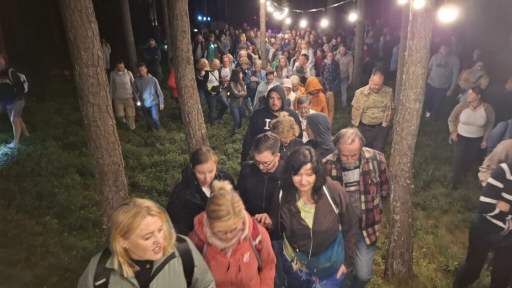 Grupa uczestników spaceruje i stoi między drzewami podczas nocnego zwiedzania; w tle widać kolejnych zwiedzających oraz oświetlenie wydarzenia (kliknięcie powiększy zdjęcie).