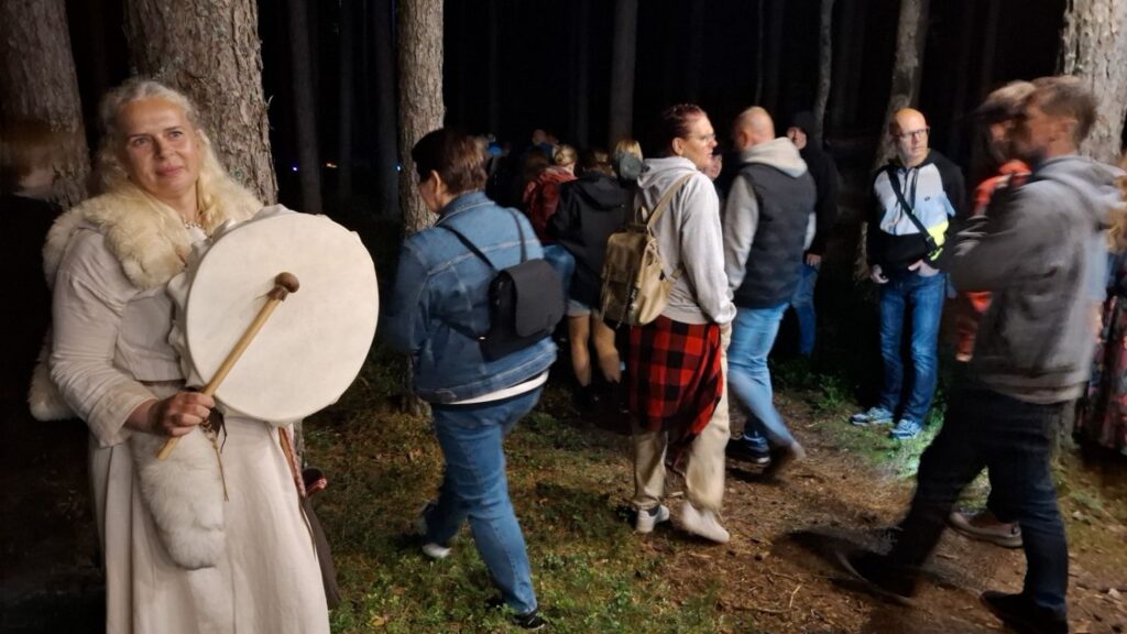 Kobieta w jasnym stroju trzyma duży okrągły bęben; wokół niej wśród drzew stoją uczestnicy nocnego zwiedzania (kliknięcie powiększy zdjęcie).