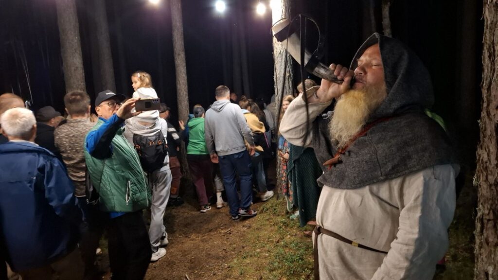 Uczestnicy wydarzenia przemieszczają się między drzewami; na pierwszym planie brodaty mężczyzna w stroju inspirowanym ubiorem historycznym gra na instrumencie - na rogu (kliknięcie powiększy zdjęcie).