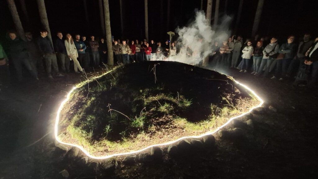 Kamienny krąg wyznaczony jasną linią światła otacza zagłębienie, z którego unosi się dym. Wokół instalacji stoją uczestnicy wydarzenia (kliknięcie powiększy zdjęcie).