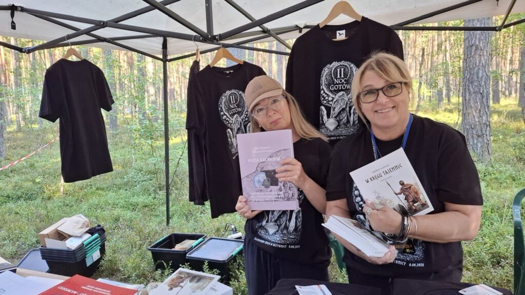 Trzy osoby stoją przy stoisku muzealnym w lesie; za nimi wiszą czarne koszulki z grafiką wydarzenia, a na stole i w ich rękach znajdują się publikacje muzealne (kliknięcie powiększy zdjęcie).