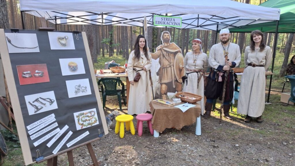 Pracownicy muzeum stoją przy stoisku edukacyjnym w lesie; obok znajduje się plansza z ilustracjami i opisami oraz stół z materiałami i eksponatami (kliknięcie powiększy zdjęcie).