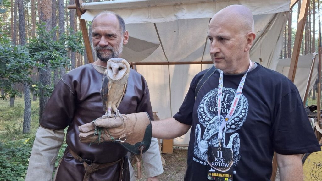 Dwóch mężczyzn prezentuje podczas wydarzenia sowę; jeden z nich trzyma ptaka w skórzanej rękawicy, a obaj mają identyfikatory i koszulki z grafiką wydarzenia (kliknięcie powiększy zdjęcie).
