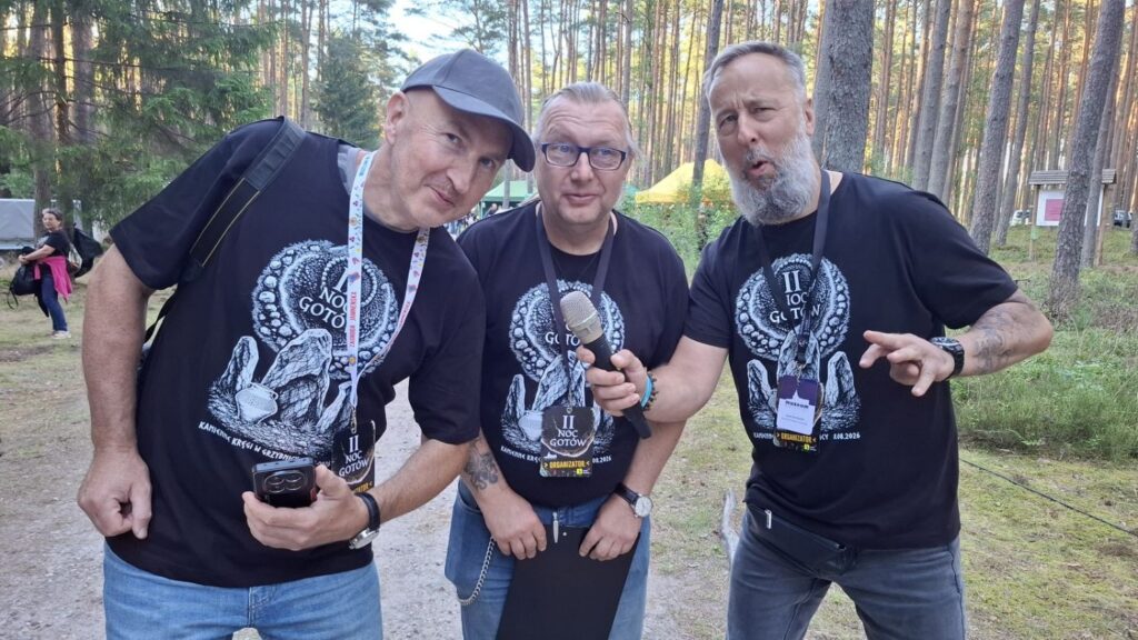 Trzech pracowników muzeum pozuje do zdjęcia w lesie; wszyscy mają czarne koszulki z grafiką wydarzenia i identyfikatory, a jeden z mężczyzn trzyma mikrofon (kliknięcie powiększy zdjęcie).