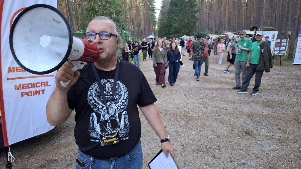 Pracownik muzeum mówi przez megafon do uczestników wydarzenia; za nim widać leśną drogę, grupę ludzi oraz oznaczenie punktu medycznego (kliknięcie powiększy zdjęcie).