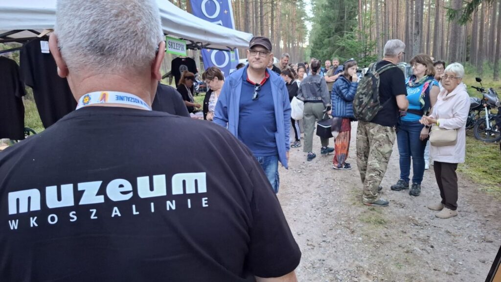 Pracownik muzeum stoi tyłem do aparatu; na jego czarnej koszulce widoczny jest napis „muzeum w Koszalinie”, a przed nim na leśnej drodze gromadzą się uczestnicy wydarzenia (kliknięcie powiększy zdjęcie).