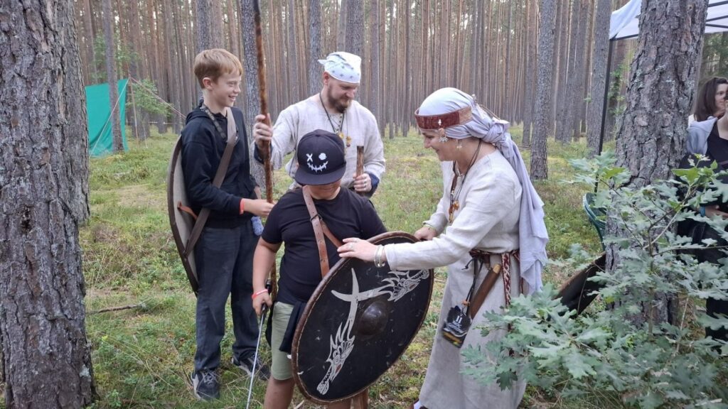 Uczestnicy wydarzenia oglądają pokaz w lesie; kobieta w stroju historycznym trzyma okrągłą tarczę, a obok stoją mężczyźni obserwujący prezentację (kliknięcie powiększy zdjęcie).